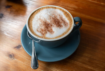 A cup of coffee with a spoon in it sits on a wooden table. The coffee is topped with whipped cream and has a sprinkle of cinnamon on top. Concept of comfort and relaxation