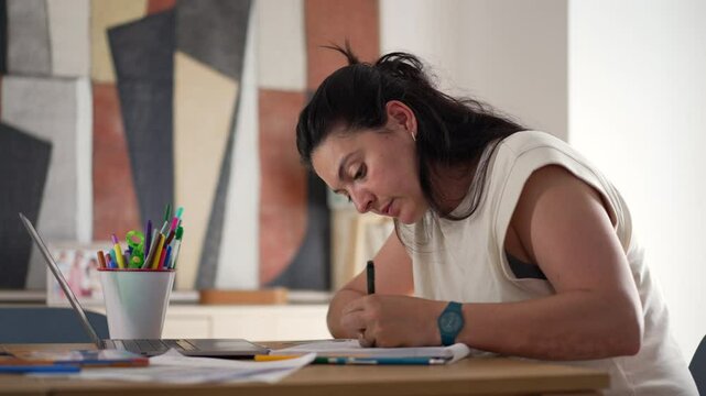 Woman studying at home desk, taking notes while following online class on laptop, focused on learning, with art supplies, paper, and books scattered around modern living room