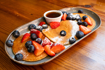 A plate of pancakes with blueberries and strawberries on top. The plate is on a wooden table