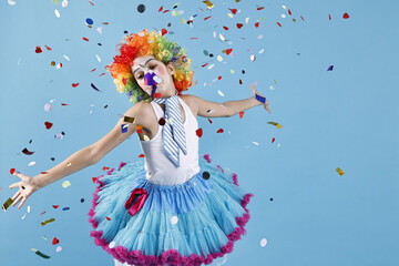 Happy girl dressed like clown under falling confetti on light blue background. Surprise party