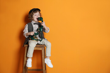Cute little boy with old telephone on stool against orange background, space for text