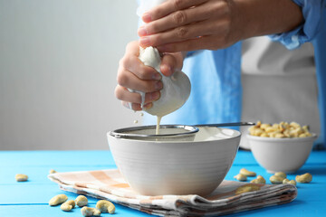 Woman straining cashew milk into bowl at light blue wooden table with nuts indoors, closeup