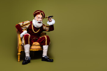 Senior man dressed as a nobleman sitting on a golden chair looking through a spyglass with a contrasting khaki background