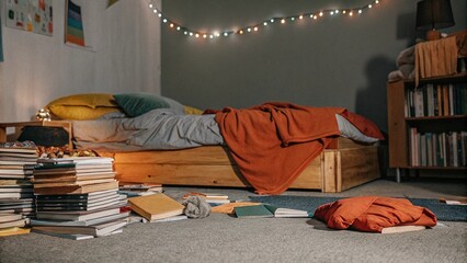 A relaxed bedroom featuring colorful bedding, stacks of books, fairy lights, and natural light streaming through wooden windows, creating a lived-in feel. Cluttercore