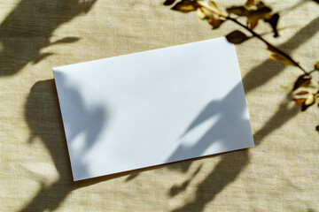 Blank white rectangular paper card on natural linen tablecloth background with shadows of leaves. Top view, flat lay. Mockup template