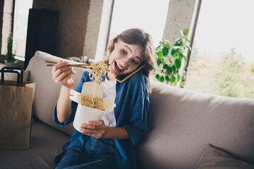 Young woman enjoying noodles while multitasking on call in cozy apartment living room