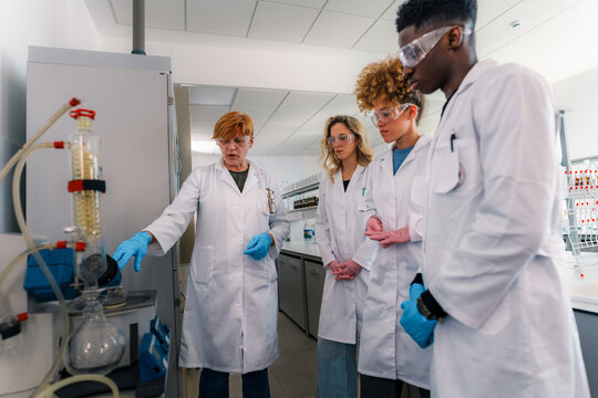 Chemistry professor wearing lab coat and gloves pointing at distillation apparatus while explaining experiment to diverse group of students in university laboratory
