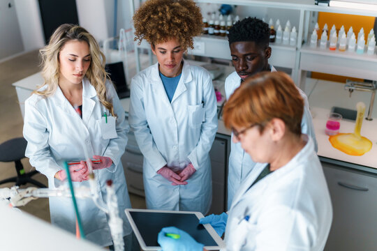 Multi ethnic university students and their professor are working together in a modern chemistry laboratory, analyzing data on a tablet and discussing their research findings