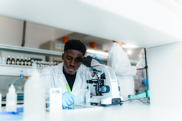 Young male scientist wearing lab coat and gloves using microscope and taking notes during...