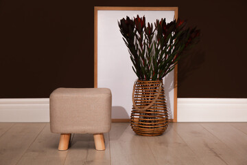 Stylish beige pouf, poster and decorative flowers near brown wall in room