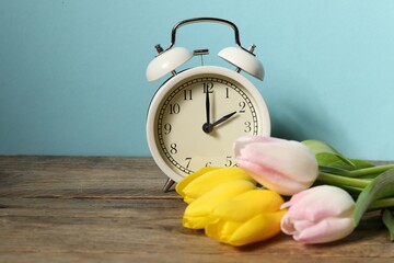 Spring time. Alarm clock and beautiful tulips on wooden table against light blue background, space...