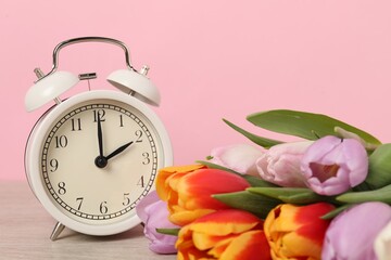 Spring time. Alarm clock and beautiful tulips on wooden table against pink background, closeup