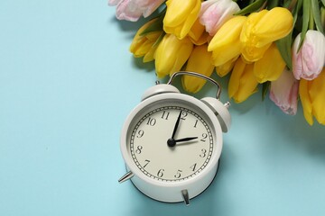 Spring time. Alarm clock and beautiful tulips on light blue background, above view. Space for text