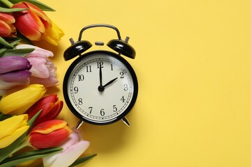 Spring time. Alarm clock and beautiful tulips on yellow background, flat lay. Space for text