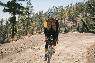 A cyclist on a gravel road in a mountainous region. He's wearing a helmet, sunglasses, and a vest....