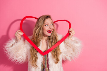 Young woman in stylish fur coat holding a heart-shaped decoration on a pink background with a...