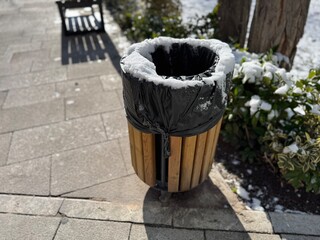 Brown wooden garbage life and snow. Modern a wooden trash can stands in a green park on the sidewalk. Outdoor wooden trash bin in park environment. Eco-friendly trash can. Close-up.
