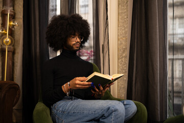 Young man sporting afro hair, wearing black turtleneck, glasses, painted nails, sitting on green...
