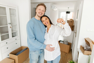 Happy European husband and pregnant wife embracing and showing key from new house, celebrating moving and smiling at camera