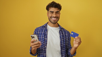 Smiling young hispanic man with beard holding smartphone and credit card against isolated yellow...