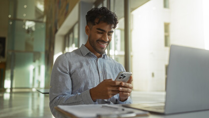 Man smiling while using smartphone in clinic with laptop on desk, sunlight streaming through...