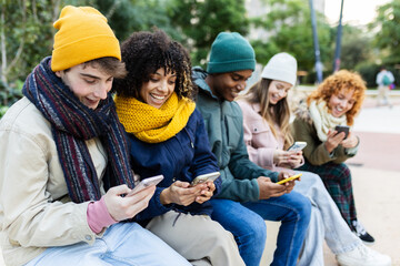 Young group of diverse teen friends using mobile phones sitting on bench. Multiracial Gen Z student people in warm clothes browsing social media and connecting online. Youth and technology concept
