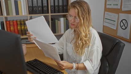 Young woman reading documents at office desk with computer, bookshelf, and bulletin board in the...