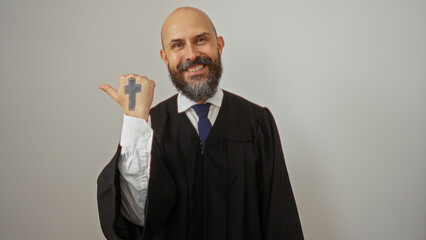 Hispanic man in a black robe, bald with a beard, isolated on a white background, smiling and pointing to the side with a thumb tattoo of a cross.