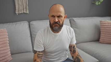 Fototapeta premium Hispanic man with a beard sitting in an apartment living room showcasing his tattooed arms against a grey couch background