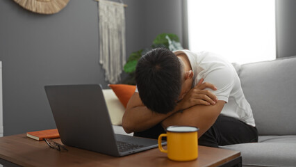 Young man indoors in a living room with a laptop and coffee mug, looking frustrated or stressed...