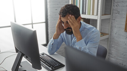 Young man with a beard looking stressed in an office setting with his hands on his head in front of...