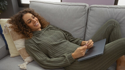 Woman relaxing on sofa with laptop in cozy living room, wearing comfortable green clothing and smiling, showcasing a serene indoor setting perfect for unwinding.