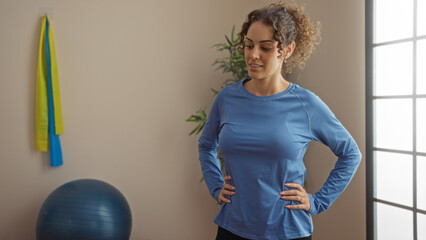 Woman smiling confidently in a gym wearing a blue shirt with exercise ball and bands visible in the...
