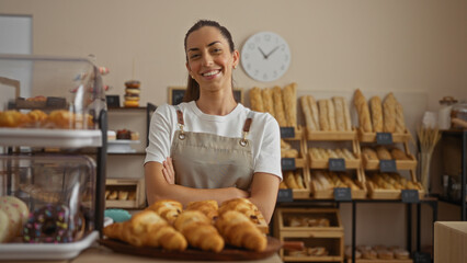 Young woman with crossed arms standing confidently inside a bakery shop, surrounded by fresh bread and pastries, smiling in front of wooden shelves filled with loaves.