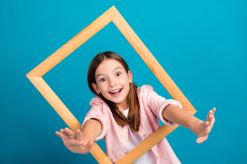Cheerful young girl posing in a wooden square frame with a vibrant blue backdrop, expressing happiness and fun in a casual pink outfit.