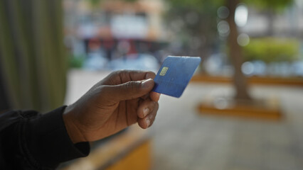 Young man holds credit card outdoors in urban park, showcasing modern banking and digital payment methods in a lively city environment with blurred street background.