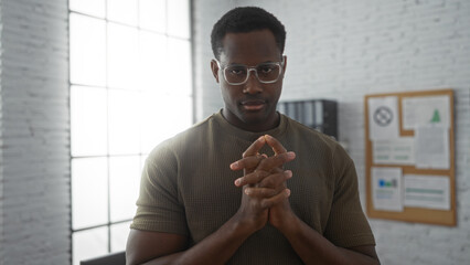Young man with serious expression and hands together standing in an office with bright light,...
