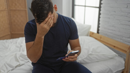 Young man with credit card and phone in hand sitting on bed in bedroom looking worried showing...
