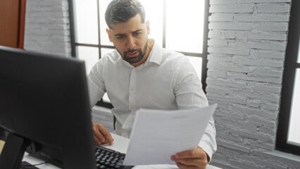 Hispanic man in office wearing white shirt intensely reading a document at his desk with computer...