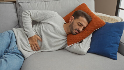 Fototapeta premium Young man with stomach pain resting on a sofa at home, displaying discomfort in a cozy living room setting with soft pillows.
