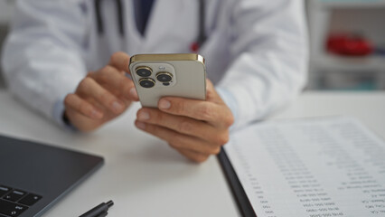 Doctor in clinic uses smartphone hands-on resting desk with laptop nearby, focusing on patient data, exemplifying modern healthcare technology.