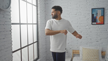 Obraz premium Young man stretches in a bedroom with sunlight streaming through a large window, surrounded by white brick walls and stylish decor