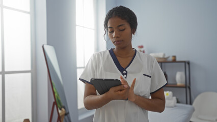Young woman in a spa room holding a tablet, wearing a uniform, with a serene expression in a well-lit, modern wellness center, showcasing the tranquil beauty interior.