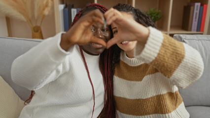 Women friends sitting together in a cozy apartment living room, sharing a heart gesture,...