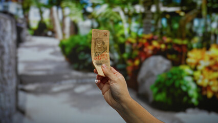 Man holding brazilian real banknote outdoors in a vibrant city park environment, highlighting urban lifestyle and financial activity.