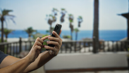 Man using smartphone outdoors, focusing on his hands with a scenic palm tree backdrop on a sunny day, showcasing a relaxed urban street environment by the ocean.