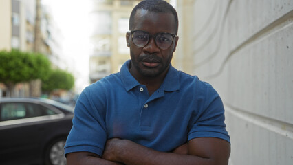 Young man standing confidently on an urban street with arms crossed, wearing glasses and a blue...