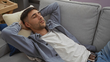 Fototapeta premium Young man relaxing on a sofa indoors with his arms behind his head, sporting casual attire, conveying a sense of comfort and tranquility in a modern home setting.