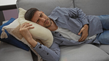 Young man experiencing stomach pain while lying on a sofa in a cozy living room, holding his abdomen with a pained expression, depicting discomfort in a home setting.
