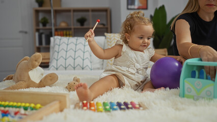 Toddler girl playing with toys on a bed in a cozy home bedroom while woman sits nearby in an indoor...